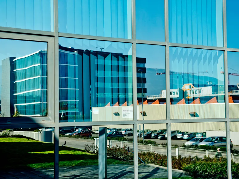 Façade vitrée d'un bâtiment moderne de la zone ZIPLO reflétant un immeuble et son parking