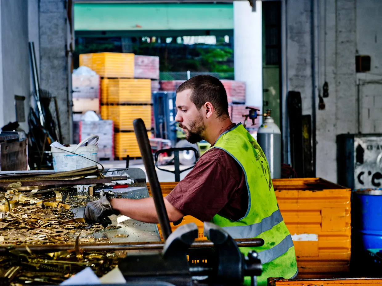 Ouvrier en gilet de sécurité travaillant sur un établi parmi des pièces métalliques, dans un atelier