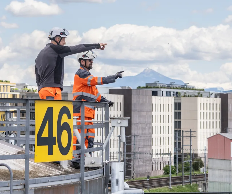 Deux ouvriers en gilet de sécurité sur une installation, désignant l'horizon industriel genevois