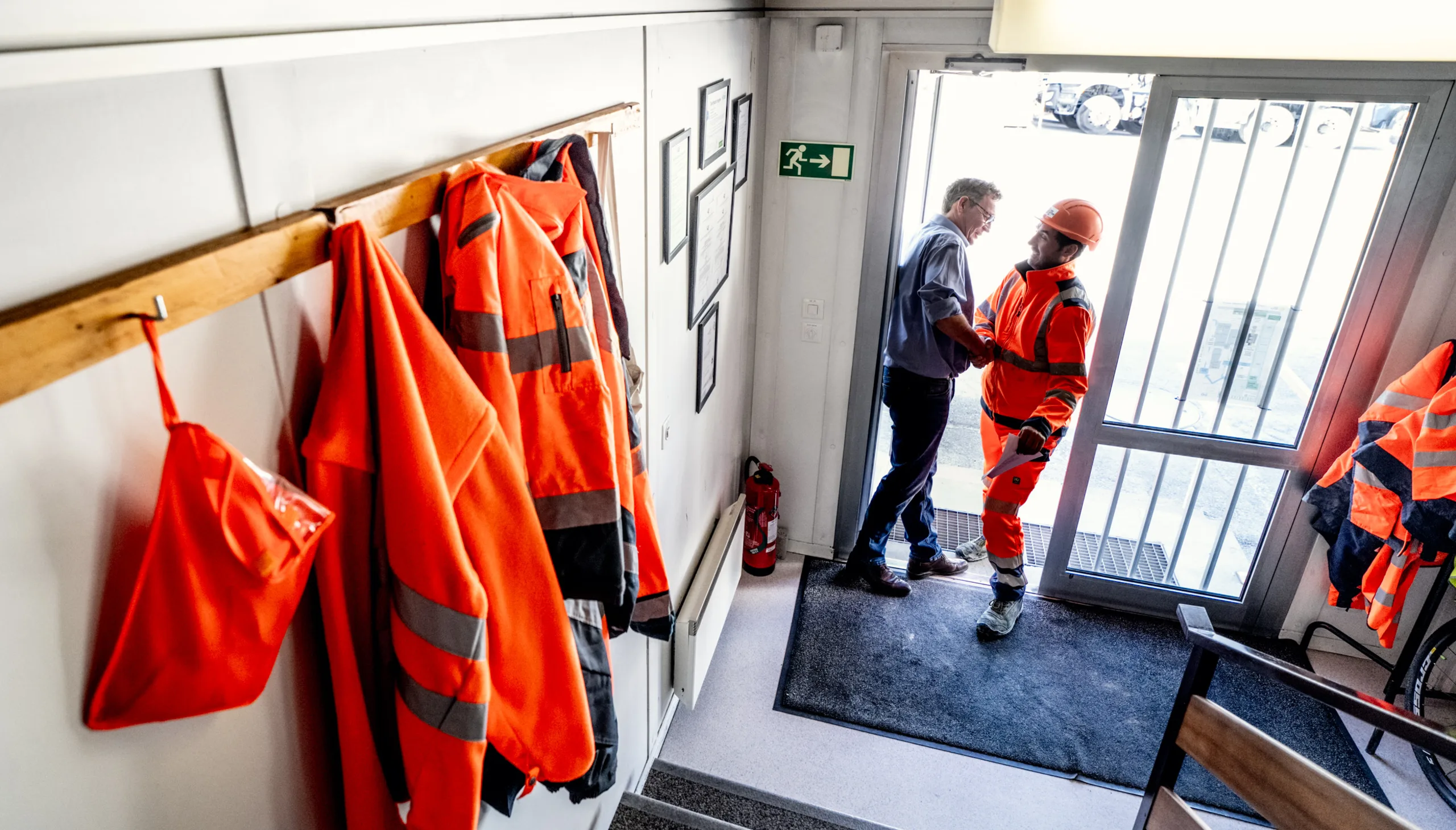Entrée d'un bâtiment de chantier : vestes de sécurité orange accrochées et ouvriers à la porte
